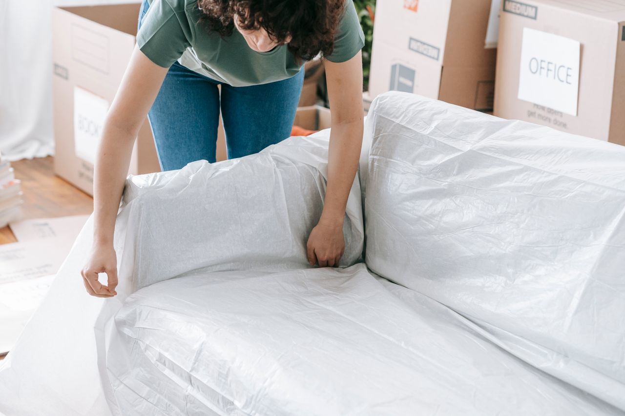 about-02 A woman carefully wraps a sofa in protective material while packing boxes in a bright room.