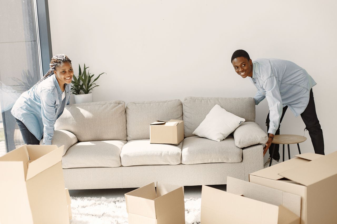 hero-img-02 A cheerful couple moving furniture in a well-lit modern living room with cardboard boxes.